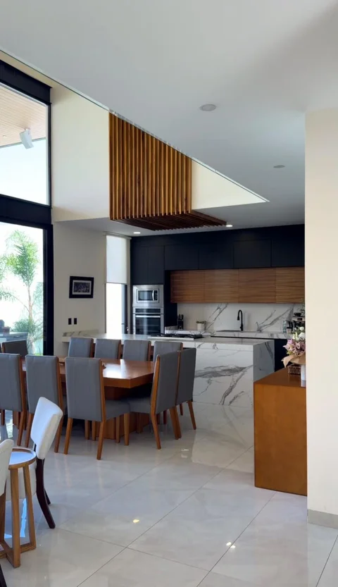 Cocina abierta con isla de cuarzo, gabinetes negros y de madera, torre de hornos y comedor